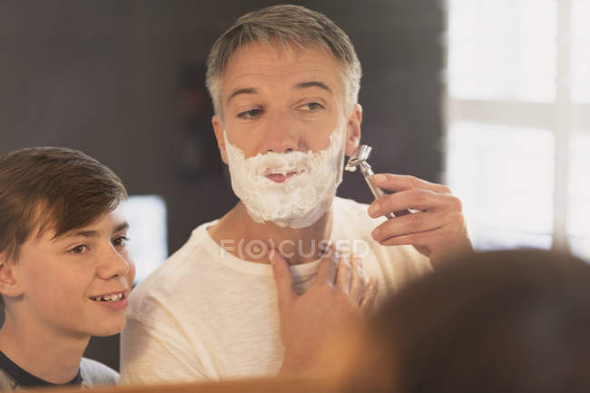 Hijo viendo padre afeitarse la cara en el espejo del baño - foto de stock
