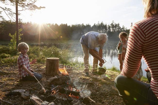 Grandparents and grandchildren at campfire at sunny lakeside in woods — Stock Photo