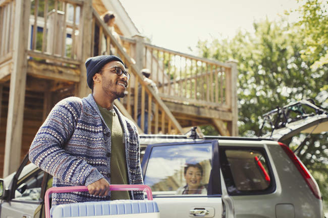 Sonriente hombre llevando refrigerador fuera del coche y cabina soleada - foto de stock