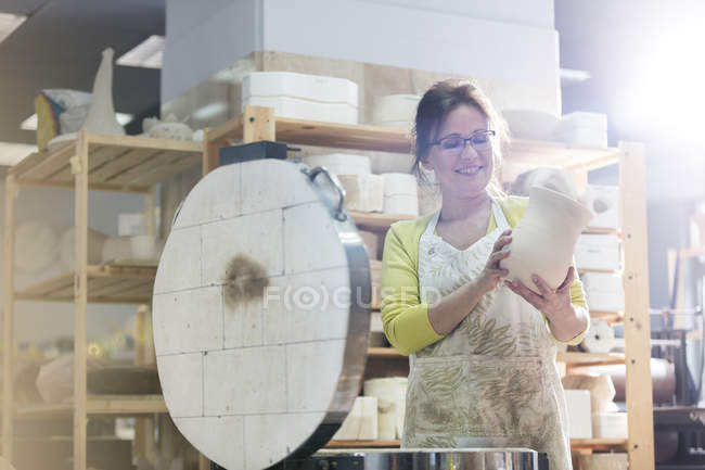Mujer madura sonriente colocando jarrón de cerámica en el horno en el estudio - foto de stock