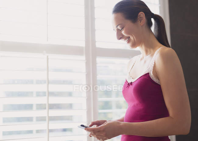 Усміхнена жінка смс з мобільним телефоном у вікні — стокове фото