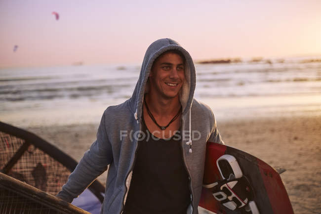 Улыбающийся мужчина в капюшоне, несущий на пляже кайтборд — стоковое фото