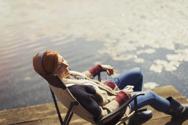 Mujer serena relajante escuchando música con auriculares y teléfono inteligente en el muelle soleado junto al lago - foto de stock