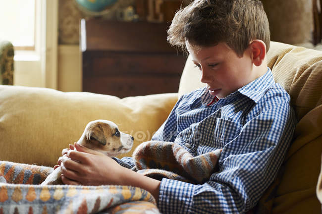 Menino segurando cachorro no colo no sofá em casa — Fotografia de Stock