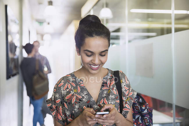 Sonriente estudiante universitaria mensajes de texto en el pasillo - foto de stock