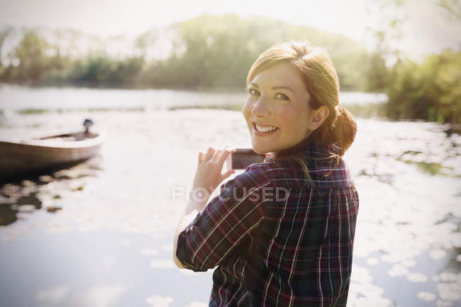 Portrait femme souriante aux cheveux roux photographiant lac ensoleillé avec téléphone appareil photo — Photo de stock