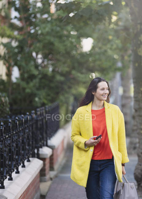 Усміхнена жінка йде з мобільним телефоном у міському парку — стокове фото