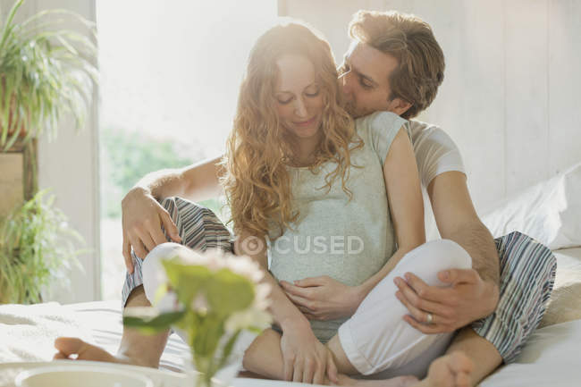 Нежная беременная пара целуется в пижаме на кровати в солнечной спальне — стоковое фото