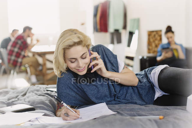 Female college student studying and talking on cell phone on bed — Stock Photo