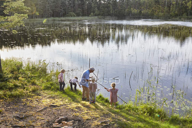 Abuelo enseñando a los nietos a pescar en el lago soleado - foto de stock