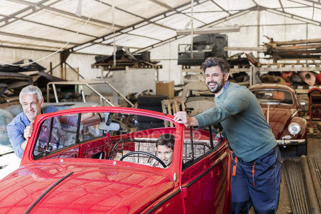Smiling mechanics pushing boy in classic car in auto repair shop — Stock Photo