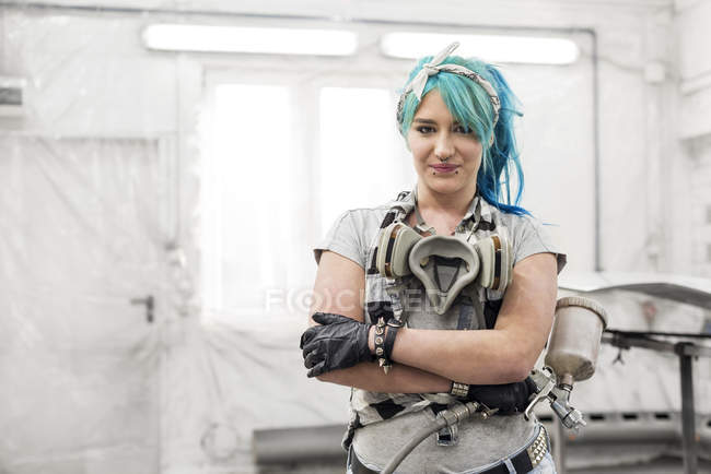 Retrato confiado joven mujer con pelo azul con pistola de pintura en taller de carrocería - foto de stock