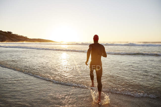 Male triathlete swimmer in wet suit running into ocean surf at sunrise — Stock Photo