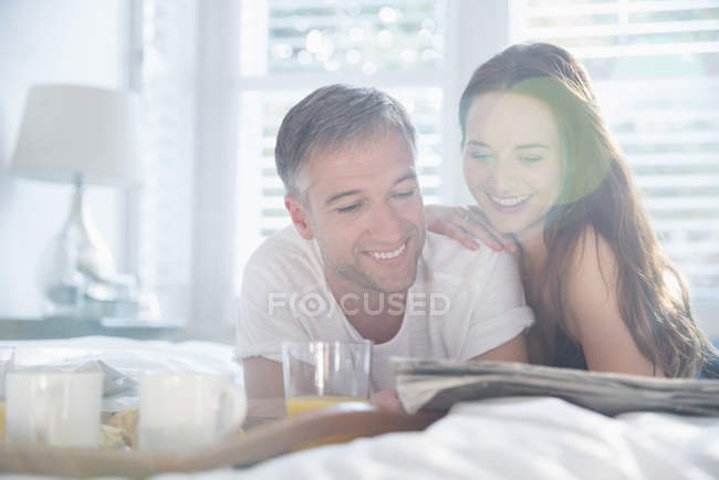 Улыбающаяся пара читает газету в солнечной спальне — стоковое фото
