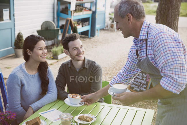 Garçom servindo cappuccinos para casal sorrindo um café ao ar livre — Fotografia de Stock