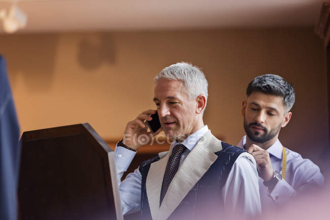 Портной примерка бизнесмена на мобильный телефон для костюма в магазине мужской одежды — стоковое фото