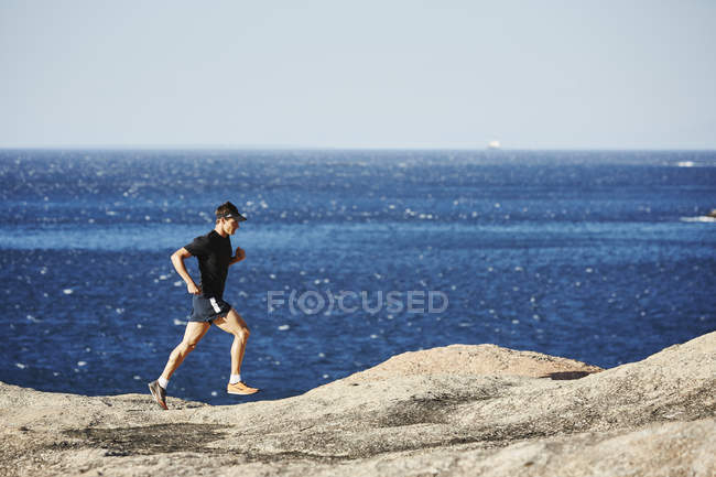 Corredor de triatleta masculino corriendo a lo largo de camino rocoso océano - foto de stock