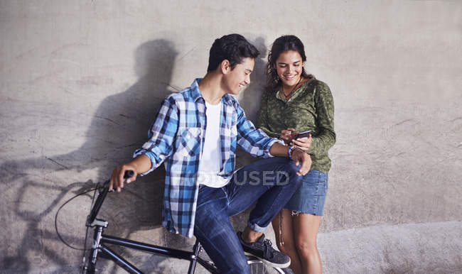 Pareja adolescente con BMX mensajes de texto en bicicleta con teléfono celular en la pared - foto de stock