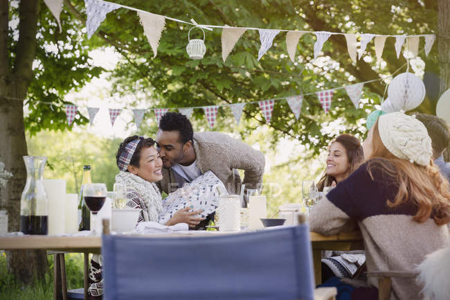 Boyfriend kissing girlfriend with birthday gift at garden party table — Stock Photo