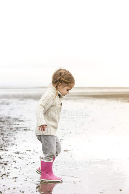 Menina em botas de chuva rosa jogando na água na praia — Fotografia de Stock