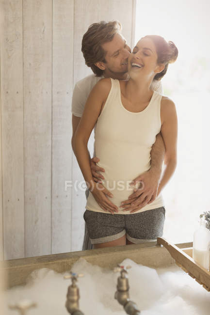 Смеющаяся нежная беременная пара целуется, готовя пенную ванну. — стоковое фото