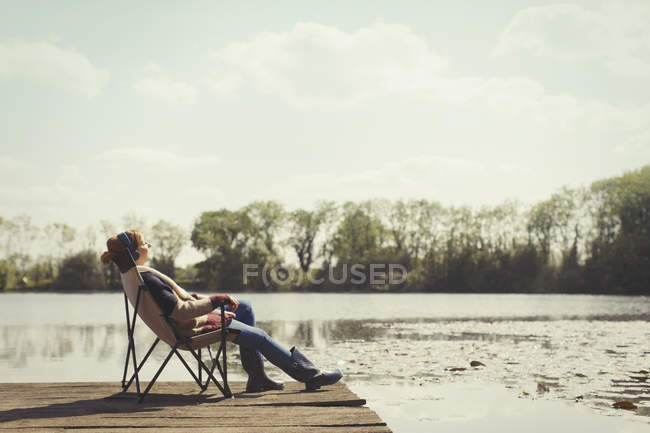 Donna serena che ascolta musica con le cuffie al molo soleggiato sul lago — Foto stock