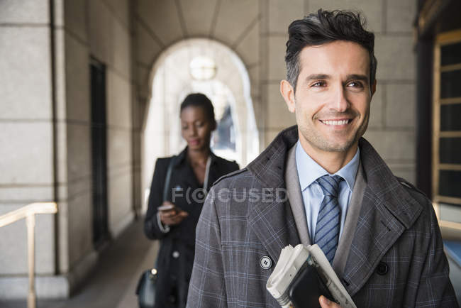 Uomo d'affari aziendale sorridente che trasporta giornali e cellulari nel chiostro — Foto stock