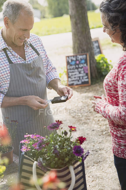 Femme avec des fleurs regardant le travailleur de pépinière d'usine utilisant la machine de carte de crédit — Photo de stock