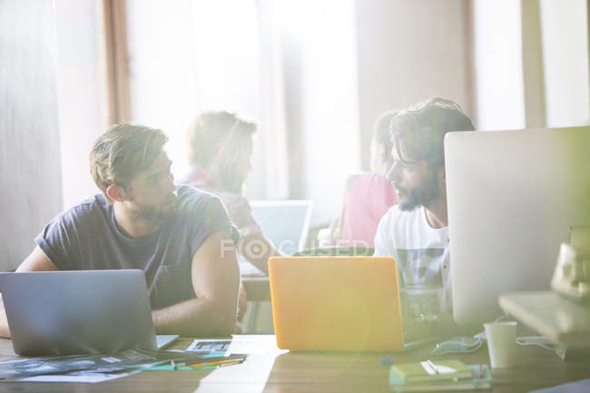 Empresarios creativos trabajando en computadoras portátiles y hablando en la oficina - foto de stock
