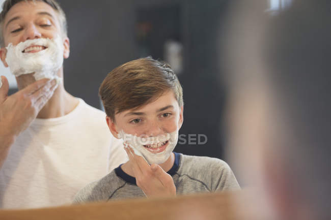 Padre viendo a su hijo fingiendo afeitarse la cara en el espejo del baño - foto de stock