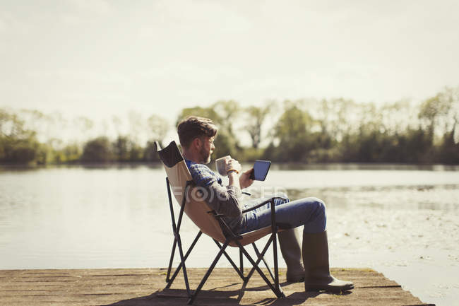 Man drinking coffee and using digital tablet on sunny lakeside dock — Stock Photo