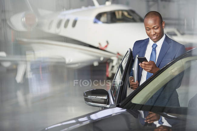 Businessman with cell phone getting into car near corporate jet in hangar — Stock Photo
