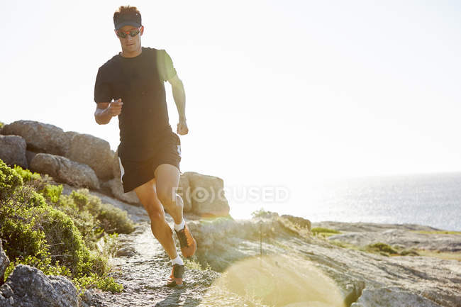 Male triathlete running on sunny rocky trail along ocean — Stock Photo