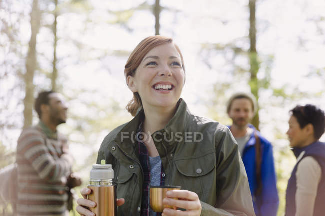 Femme souriante buvant du café dans un récipient à boisson isolé randonnée dans les bois — Photo de stock
