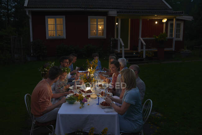 Familia disfrutando de la cena a la luz de las velas en la mesa del patio fuera de casa por la noche - foto de stock
