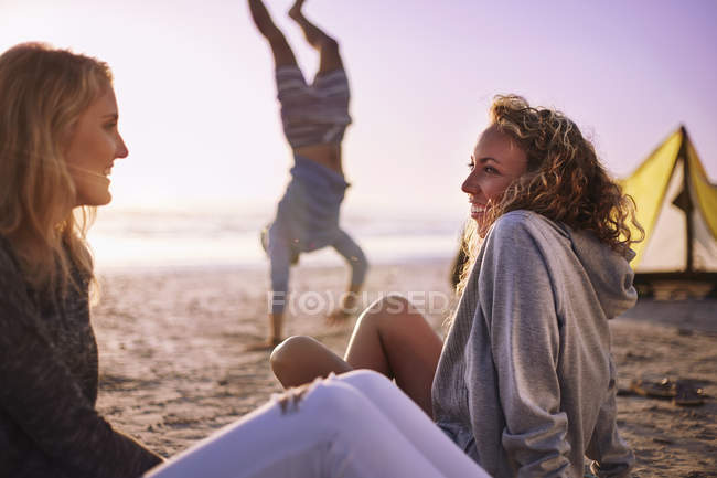 Улыбающиеся женщины разговаривают на пляже с мужчиной, делая стойку на руках на заднем плане — стоковое фото