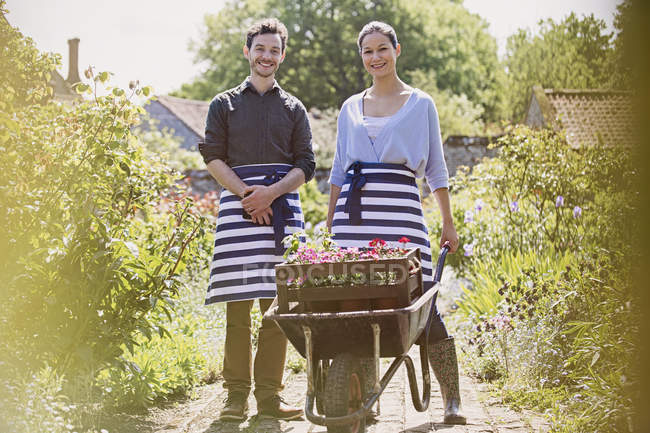 Portrait smiling plant nursery workers with wheelbarrow and flowers in sunny garden — Stock Photo