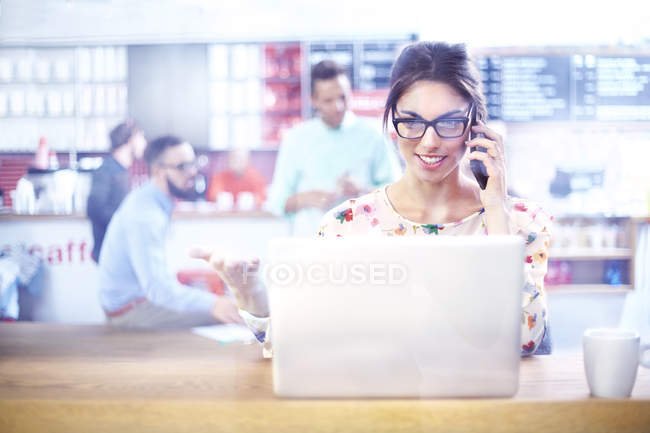 Femme d'affaires utilisant un ordinateur portable et parlant sur un téléphone portable dans un café — Photo de stock