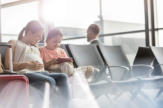 Вагітна мати і дочка використовують цифровий планшет у зоні відправлення аеропорту — стокове фото