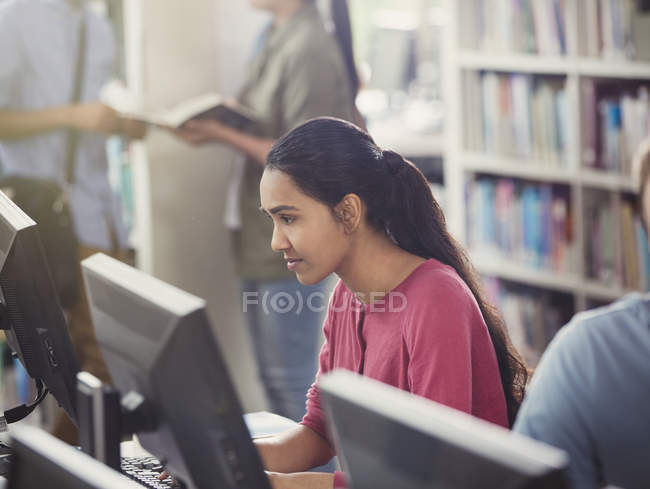 Estudiante universitaria enfocada investigando usando computadora en la biblioteca - foto de stock