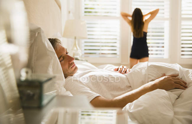 Wife stretching at morning window behind husband sleeping in bed — Stock Photo