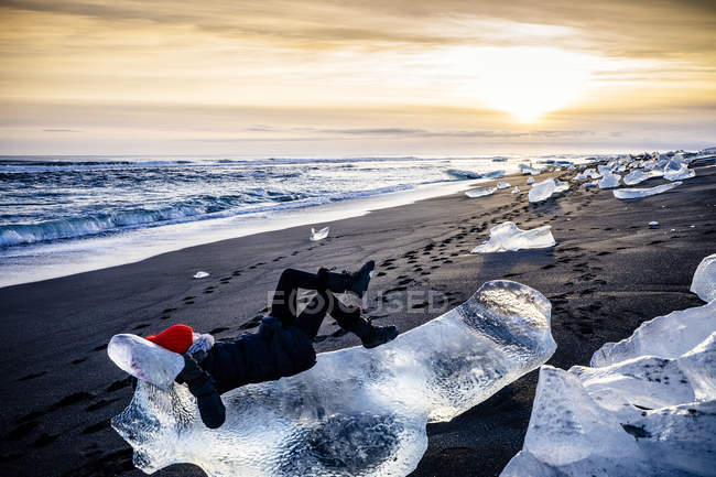 Mujer acostada en un gran trozo de hielo en la playa al atardecer, Islandia - foto de stock
