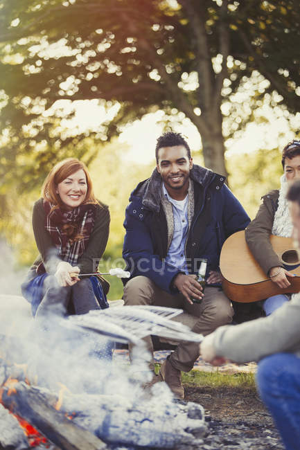 Amigos sorridentes assando marshmallows e bebendo cerveja na fogueira — Fotografia de Stock