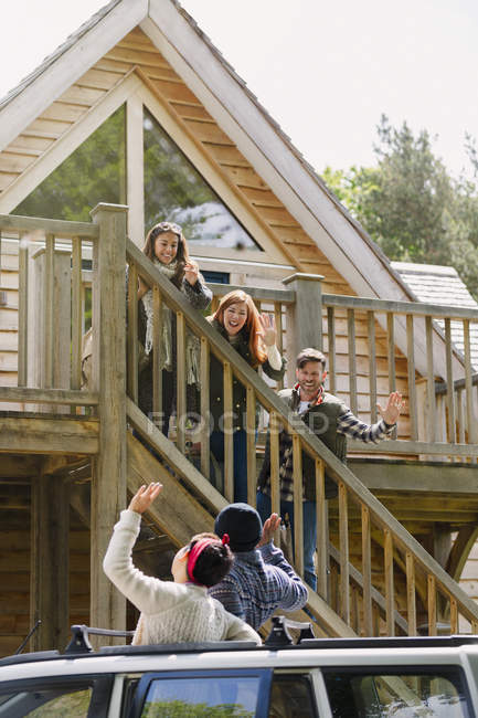 Friends on stairs waving to friends in car outside sunny cabin — Stock Photo