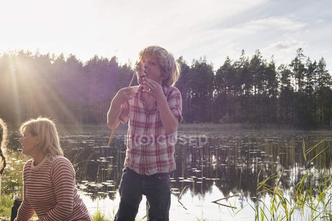 Enthusiastic boy eating roasted marshmallows at sunny lakeside in woods — Stock Photo