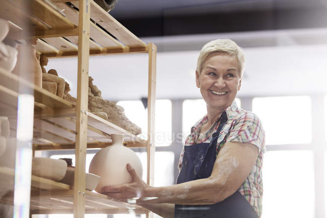 Sorrindo mulher sênior colocando vaso de cerâmica na prateleira no estúdio — Fotografia de Stock
