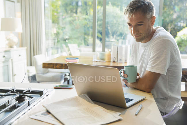 Homem bebendo café e trabalhando no laptop na cozinha — Fotografia de Stock