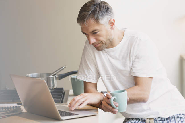 Man in pajamas drinking coffee and working at laptop in kitchen — Stock Photo
