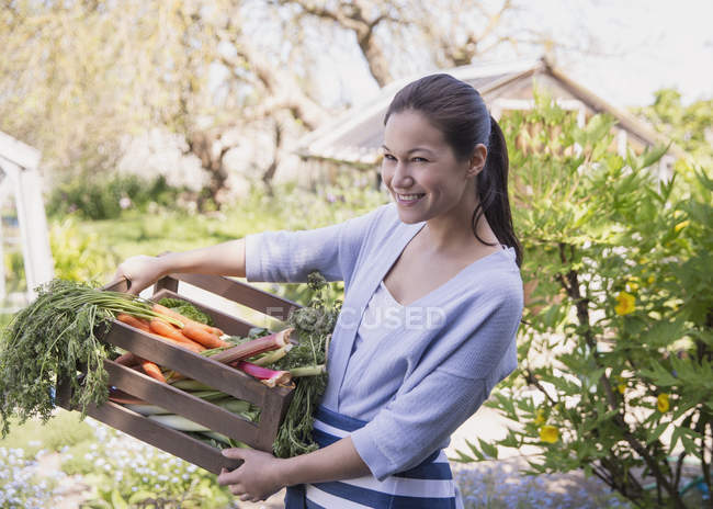 Retrato sonriente mujer sosteniendo cajón de verduras frescas cosechadas en el jardín - foto de stock