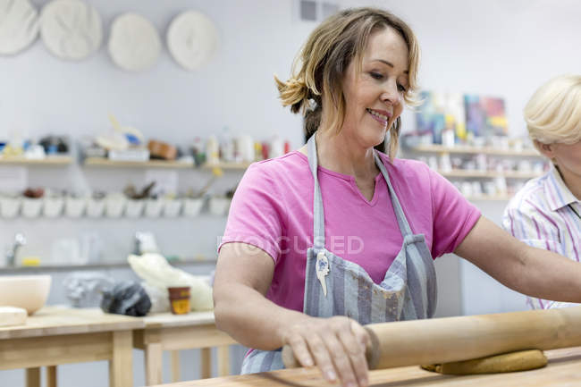 Улыбающаяся зрелая женщина катит глину со скалкой в мастерской керамики — стоковое фото